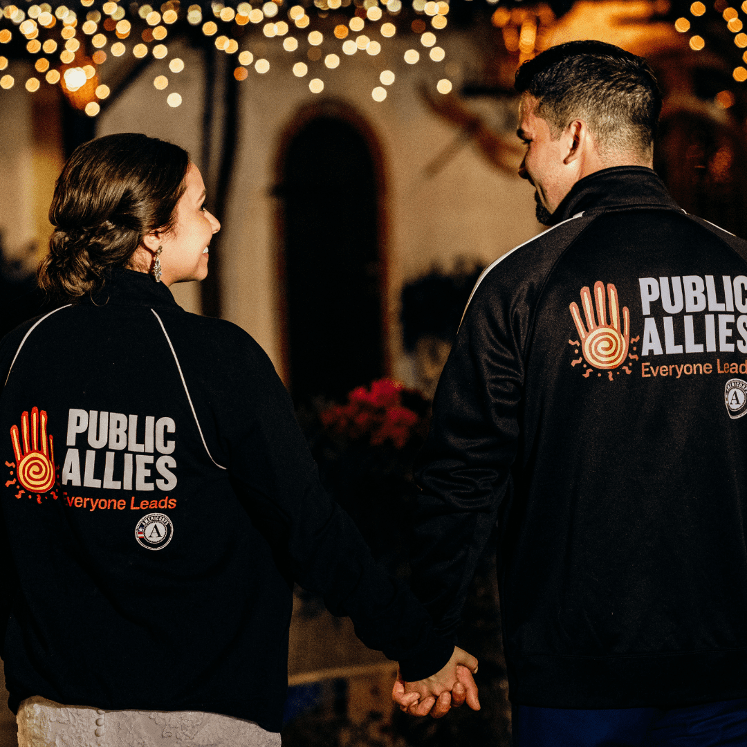 A couple stands with their backs to the camera, both wearing Public Allies branded track jackets. They are smiling at each other lovingly.